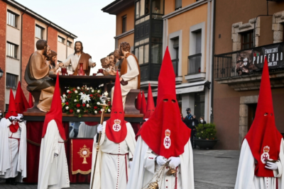 Imponente procesión de la Santa Cena en Ponferrada: el gran paso de madera tallada con Cristo y los doce apóstoles, acompañado por cofrades con túnicas y capirotes rojos de la Real Hermandad de Jesús Nazareno, avanza por la Plaza del Ayuntamiento durante la Semana Santa en El Bierzo, en Jueves Santo.