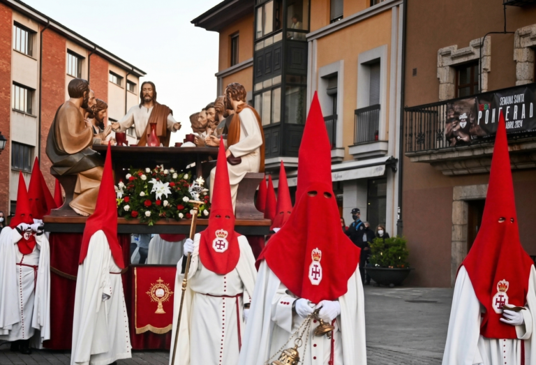 Imponente procesión de la Santa Cena en Ponferrada: el gran paso de madera tallada con Cristo y los doce apóstoles, acompañado por cofrades con túnicas y capirotes rojos de la Real Hermandad de Jesús Nazareno, avanza por la Plaza del Ayuntamiento durante la Semana Santa en El Bierzo, en Jueves Santo.