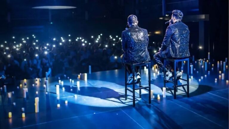 Dos cantantes de espaldas, sentados en taburetes sobre un escenario rodeado de velas, frente al público con las linternas de los móviles encendidas