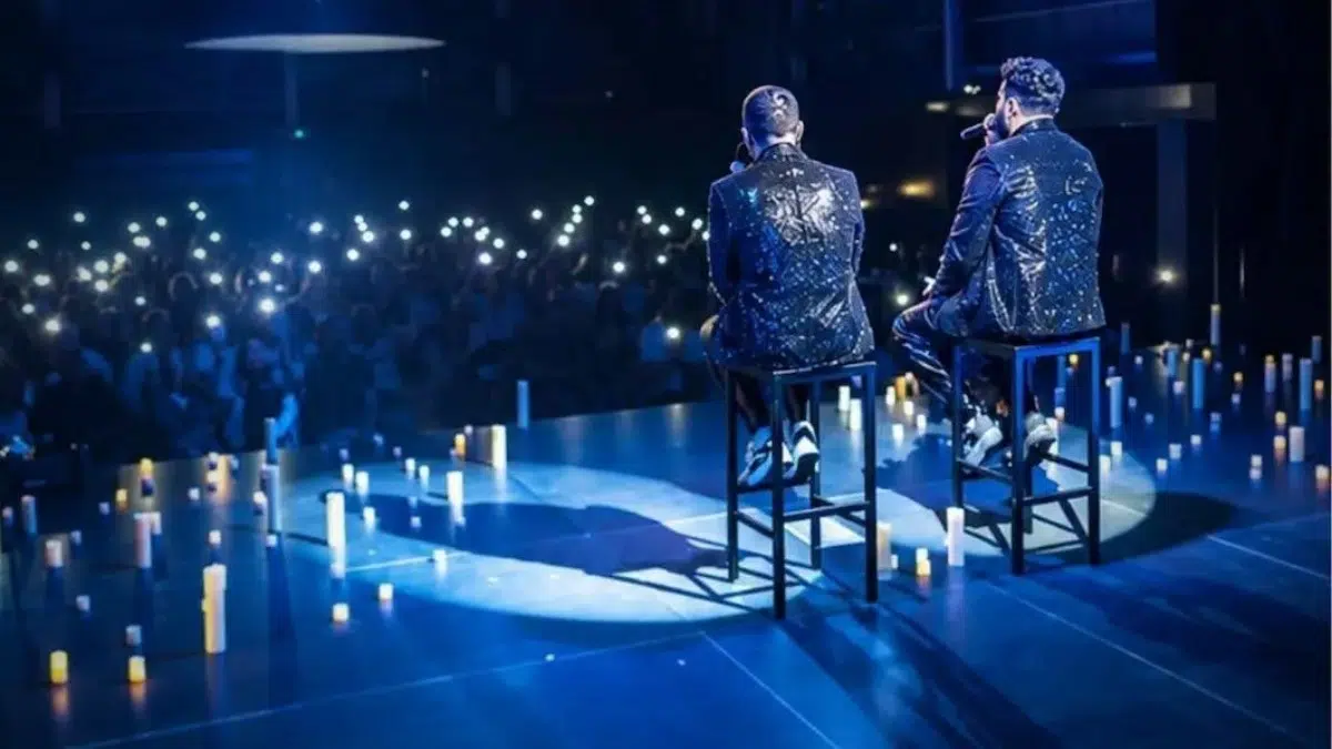 Dos cantantes de espaldas, sentados en taburetes sobre un escenario rodeado de velas, frente al público con las linternas de los móviles encendidas